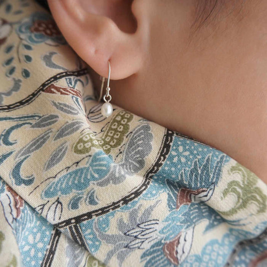 Close-up of a model's ear wearing Pearl drop French hook dangle earrings, with a colorful earth toned patterned fabric background.