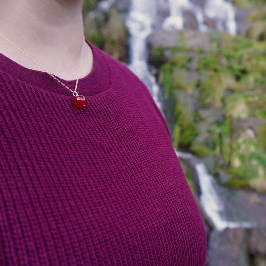 Side view of a dainty round red carnelian ball sphere pendant necklace in 14k gold plated 925 sterling silver on a 14k gold filled chain worn on a person wearing a burgundy, dark pink sweater, with a natural blurred background of a Japanese waterfall.