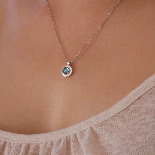 Close up view of a dainty, round Azotic blue topaz cabochon pendant sterling silver necklace, worn by a person wearing a light beige shirt.