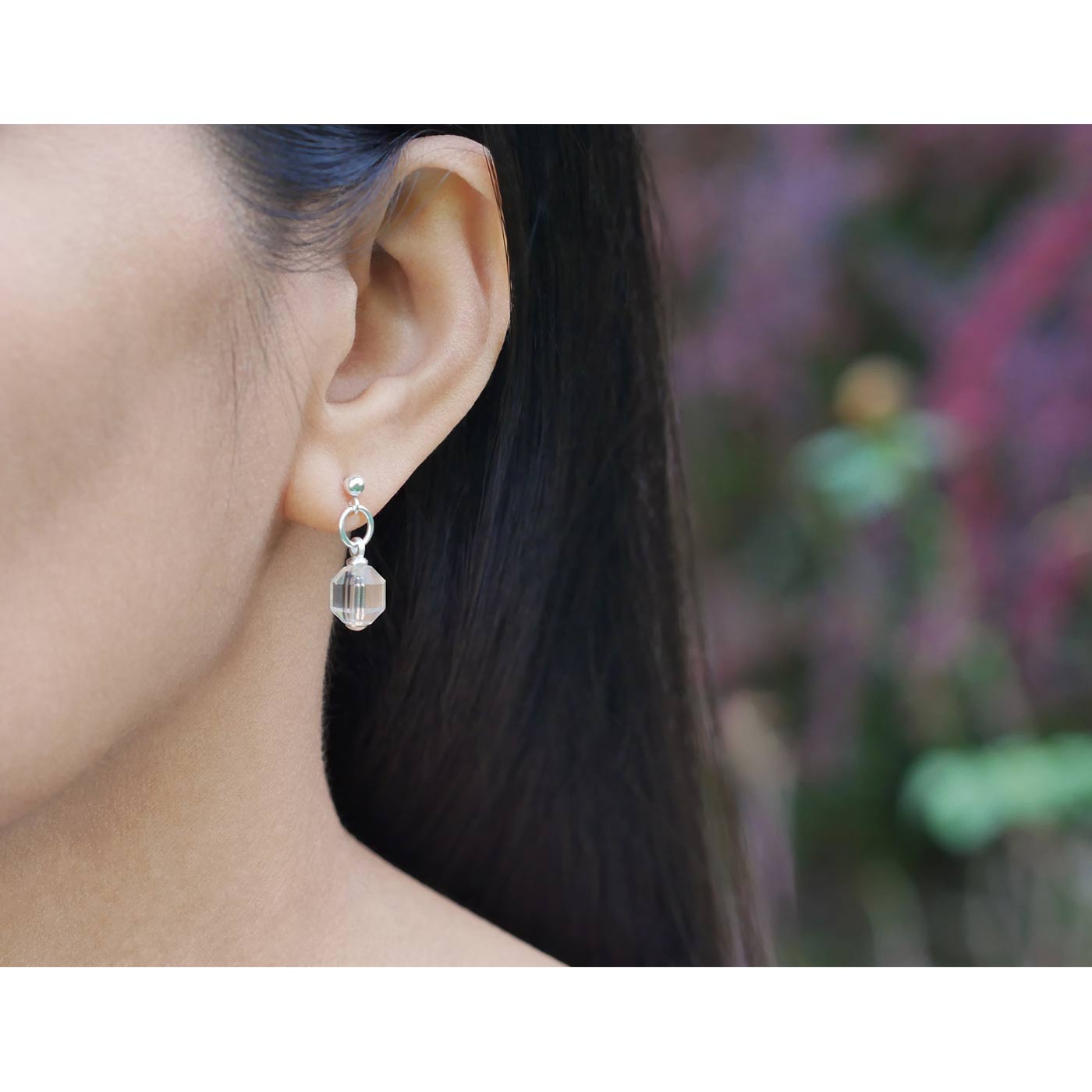 A close up of a sterling silver clear quartz crystal prism earring worn outside by a person with dark hair, against a blurred natural background with plants and flowers.