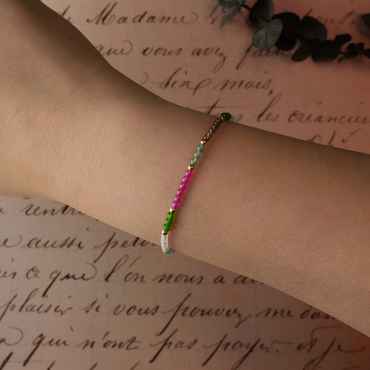 Top view of a dainty multi-colored colorful garden beaded bracelet with natural high quality AAA gemstones of Guatemalan jade, Taiwan jade, lab ruby, smokey quartz and rose quartz with 14K gold filled accent beads on a wrist with an old lettering vintage light pink beige paper and green leaves stylized in the background.