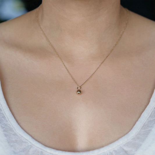 Front view of a dainty, round 14k gold filled sphere ball pendant necklace worn on a person wearing a light beige off white top.