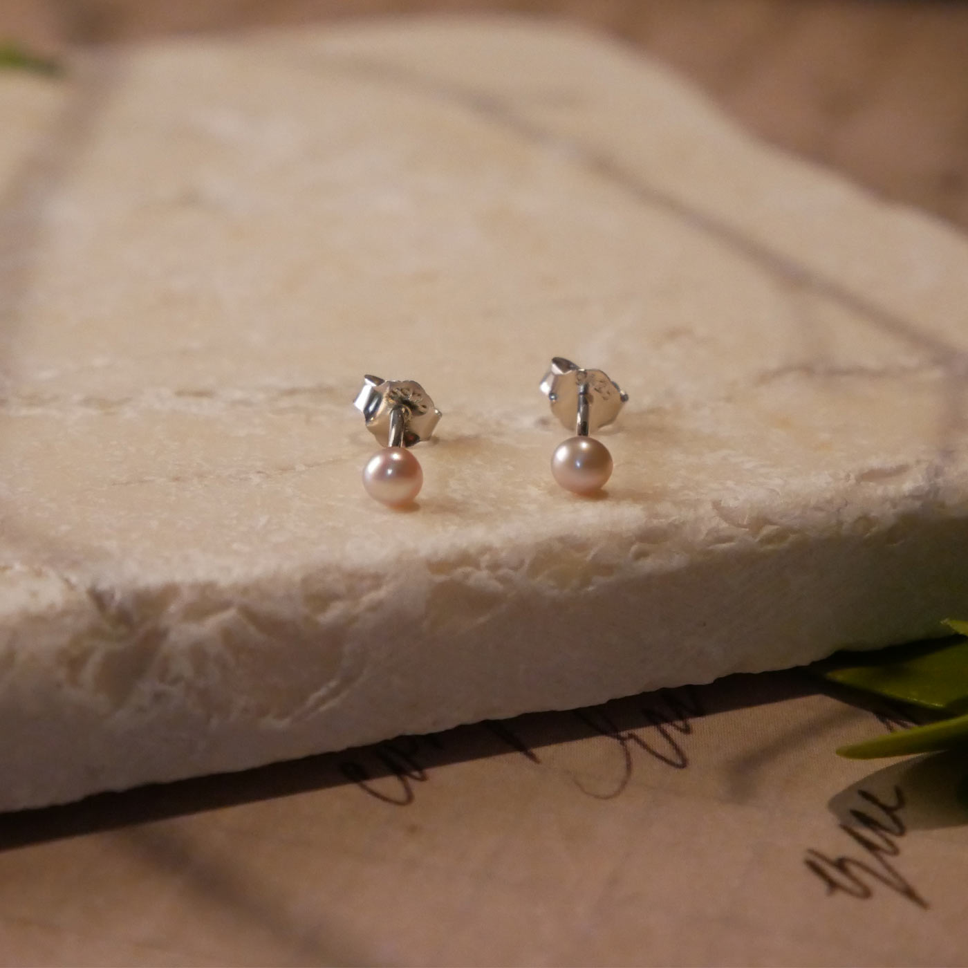 Front view of minimalist dainty pair of natural pink pearl sterling silver stud earrings on a stone surface, stylized with branch shadows, accent leaves, and on vintage paper with cursive writing.