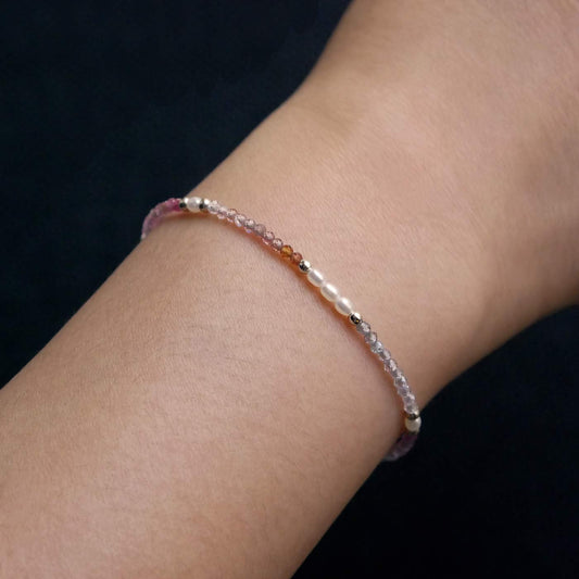 Close-up of a wrist wearing a dainty, delicate beaded bracelet with faceted colorful ombre spinel, rice pearls, and sterling silver accents. The beads are high quality AAA, the background is dark black.