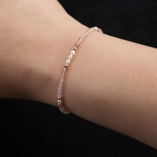 Close-up of a wrist wearing a dainty beaded bracelet with faceted strawberry quartz, rice pearls, and 14K gold filled accents. The beads are high quality AAA, the background is dark black.