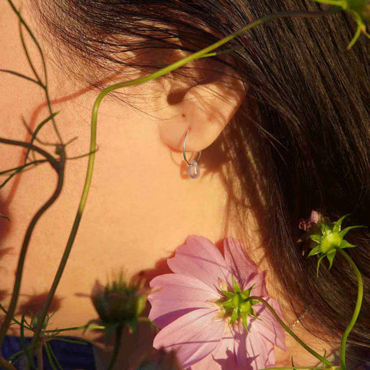 Outside close-up of a person's ear wearing a sterling silver Czech glass crystal AB aurora borealis colored drop wire hoop earring, surrounded by pink flowers and green leaves.