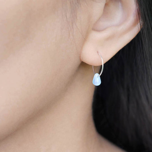 A close up of a sterling silver Czech glass crystal drop in a milky blue pink color on a wire hoop earring, worn outside by a person with dark hair.