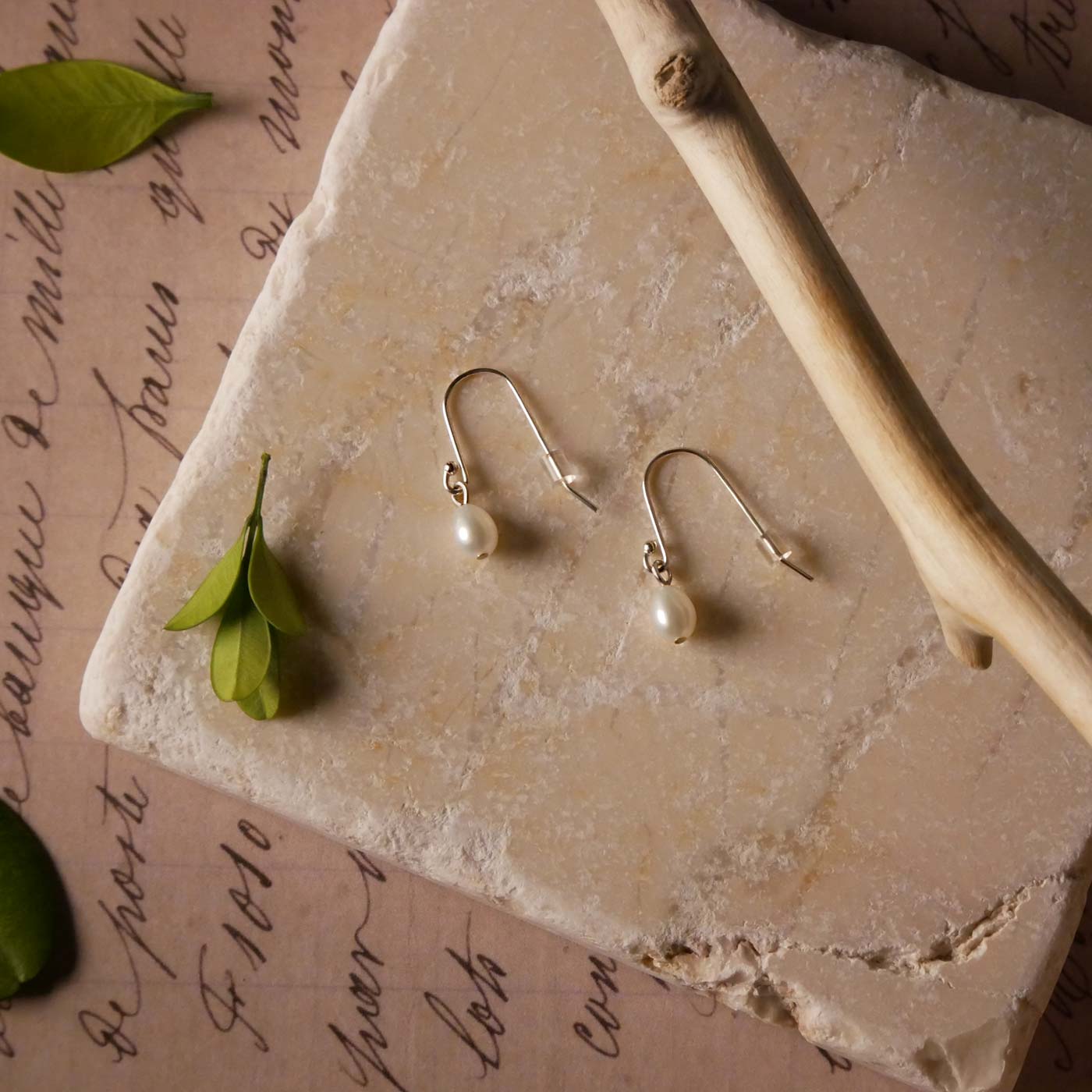 Top view of Pearl drop French hook earrings on a textured stone surface with leaves and a branch accent.