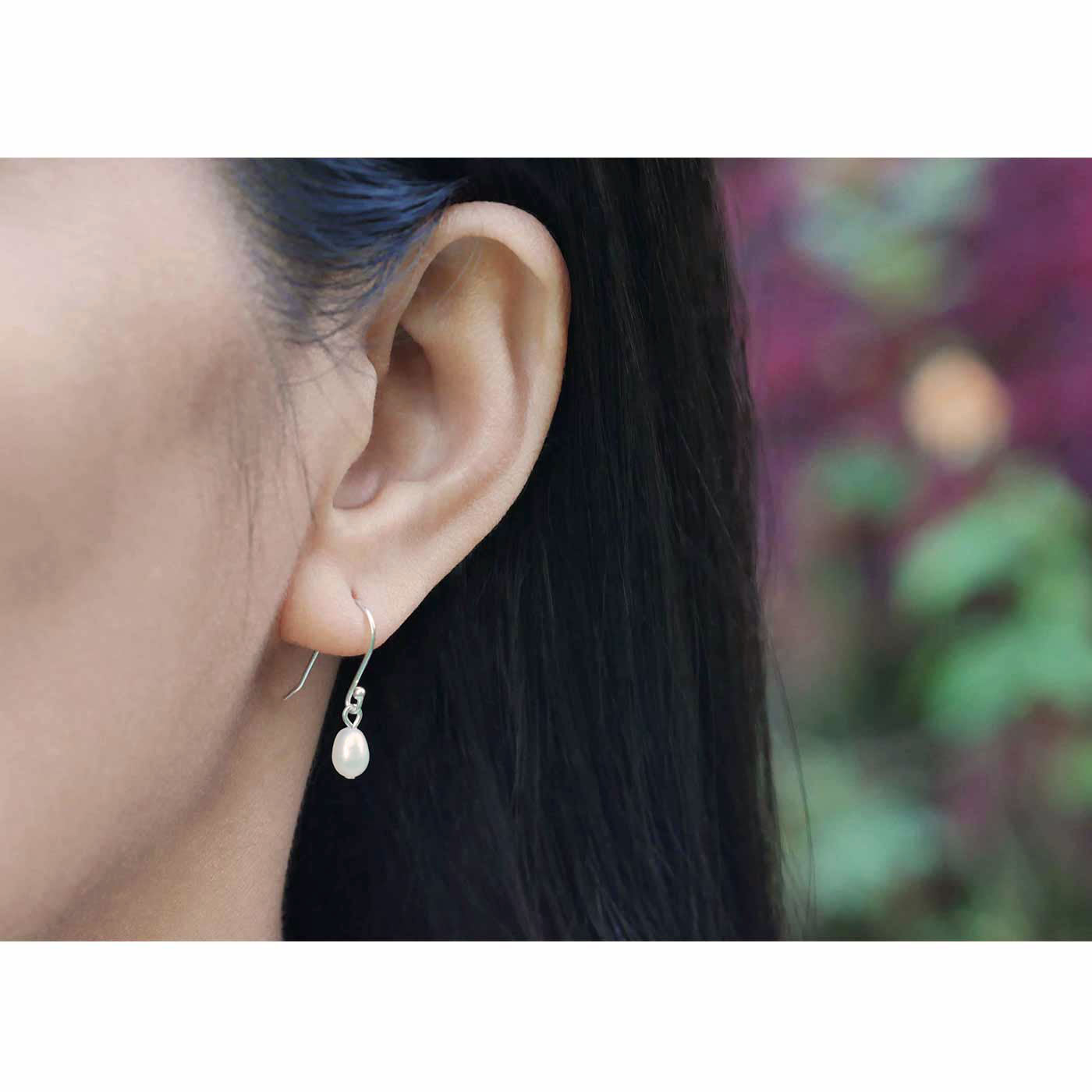 Close-up of a model's ear wearing Pearl drop French hook sterling silver earrings with a blurred flower outdoor background.