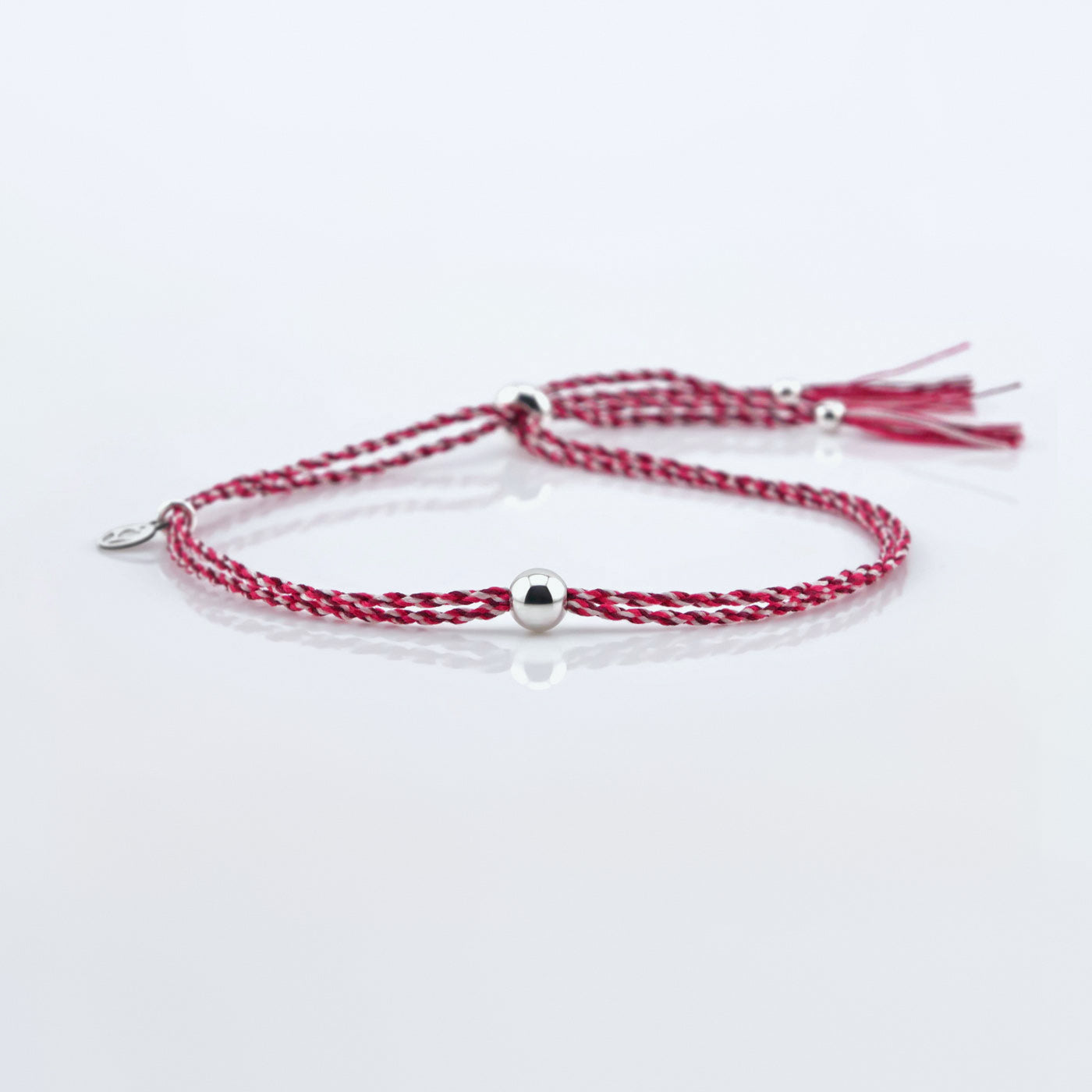 Front view of a colorful adjustable rpet cord friendship bracelet with a focal solid sterling silver ball sphere, sterling silver accents, and tassels. The twisted cord is hand spun with sakura colors of dark pink, bright pink, and white colors. The bracelet is laying flat on a reflective plain white surface background.
