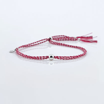 Front view of a colorful adjustable rpet cord friendship bracelet with a focal solid sterling silver ball sphere, sterling silver accents, and tassels. The twisted cord is hand spun with sakura colors of dark pink, bright pink, and white colors. The bracelet is laying flat on a reflective plain white surface background.
