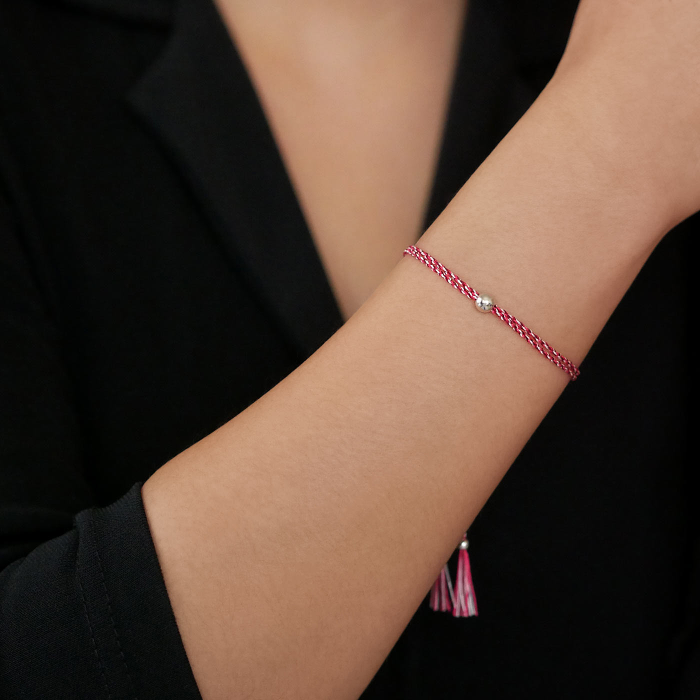 Close-up of a wrist wearing a colorful adjustable rpet cord friendship bracelet with a focal solid 925 sterling silver ball sphere, sterling silver accents and tassels. The twisted cord is hand spun with sakura inspired colors of bright pink, magenta, dark pink and white colors. The background is the person's black top.