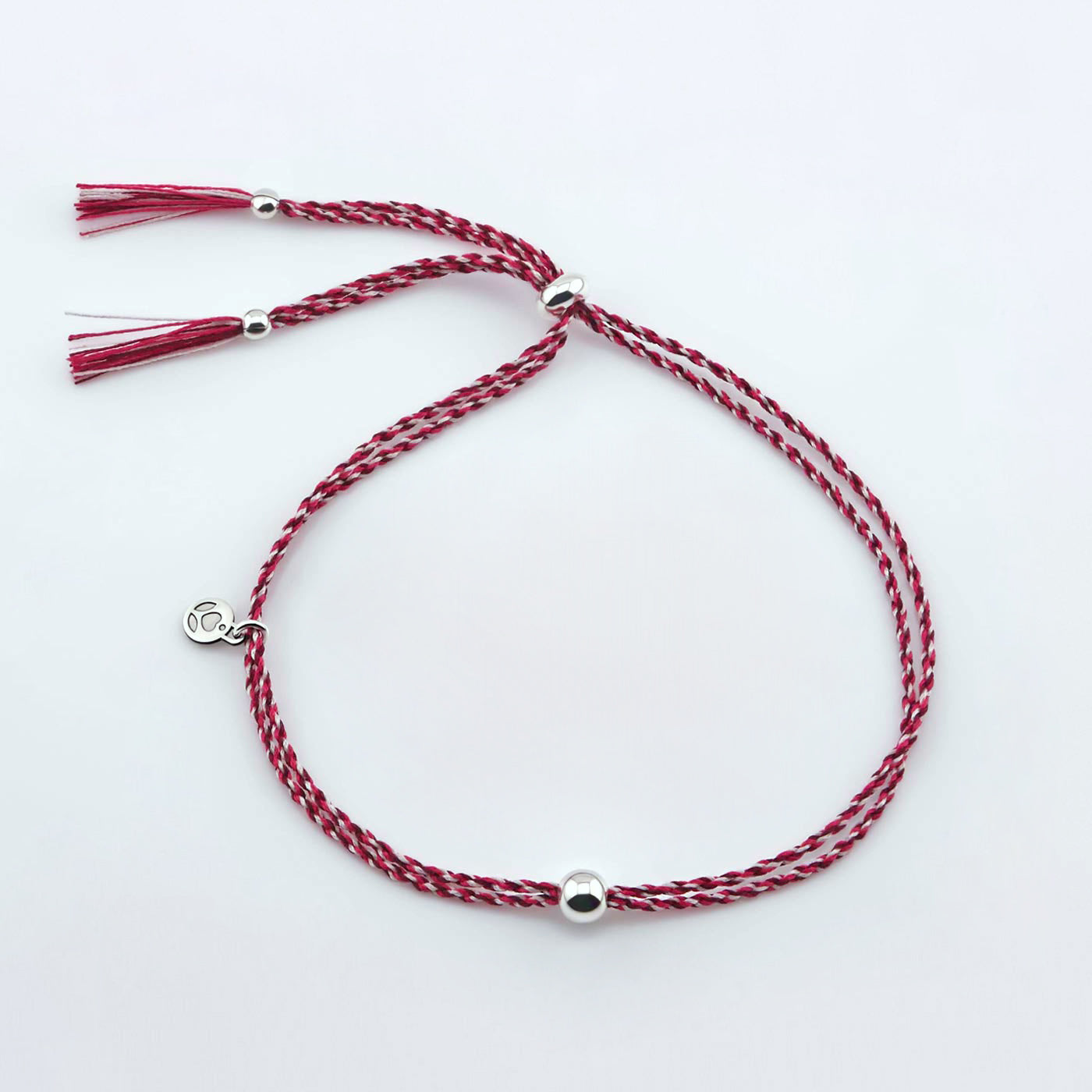 Top view of a colorful adjustable rpet cord friendship bracelet with a focal solid sterling silver ball sphere and sterling silver accents and tassels. The twisted cord is hand spun with sakura inspired colors of dark pink, bright pink, and white colors. The bracelet is laying flat on a reflective plain white surface background.
