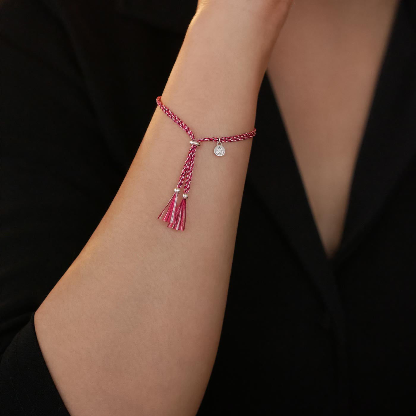 Close-up of a wrist wearing a colorful adjustable rpet cord friendship bracelet with a focal solid 925 sterling silver ball sphere, sterling silver accents, the brand logo and tassels. The twisted cord is hand spun with sakura inspired colors of bright pink, magenta, dark pink and white colors. The background is the person's black top.