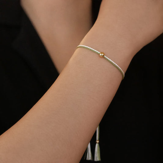 Close-up of a wrist wearing a colorful adjustable rpet cord friendship bracelet with a focal solid 14K gold ball sphere, gold accents and tassels. The twisted cord is hand spun with moss green, ivory, off white colors. The background is the person's black top.