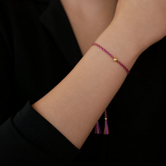 Close-up of a wrist wearing a colorful adjustable rpet cord friendship bracelet with a focal solid 14K gold ball sphere, gold accents and tassels. The twisted cord is hand spun with purple, magenta, dark pink, red and yellow colors. The background is the person's black top.