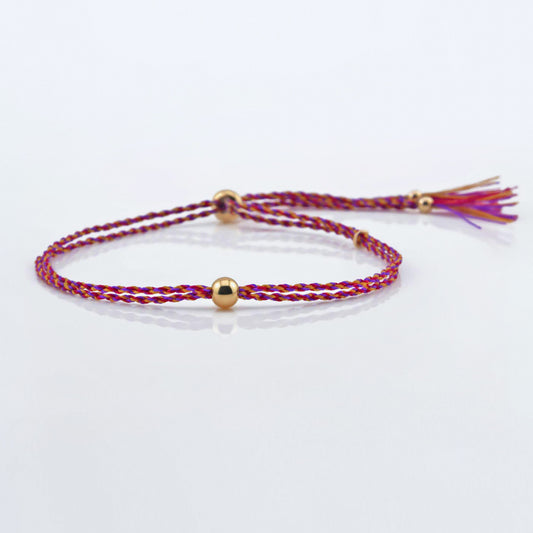 Front view of a colorful adjustable rpet cord friendship bracelet with a focal solid 14K gold ball sphere, gold accents and tassels. The twisted cord is hand spun with purple, magenta, dark pink, red and yellow colors. The bracelet is laying flat on a reflective plain white surface background.
