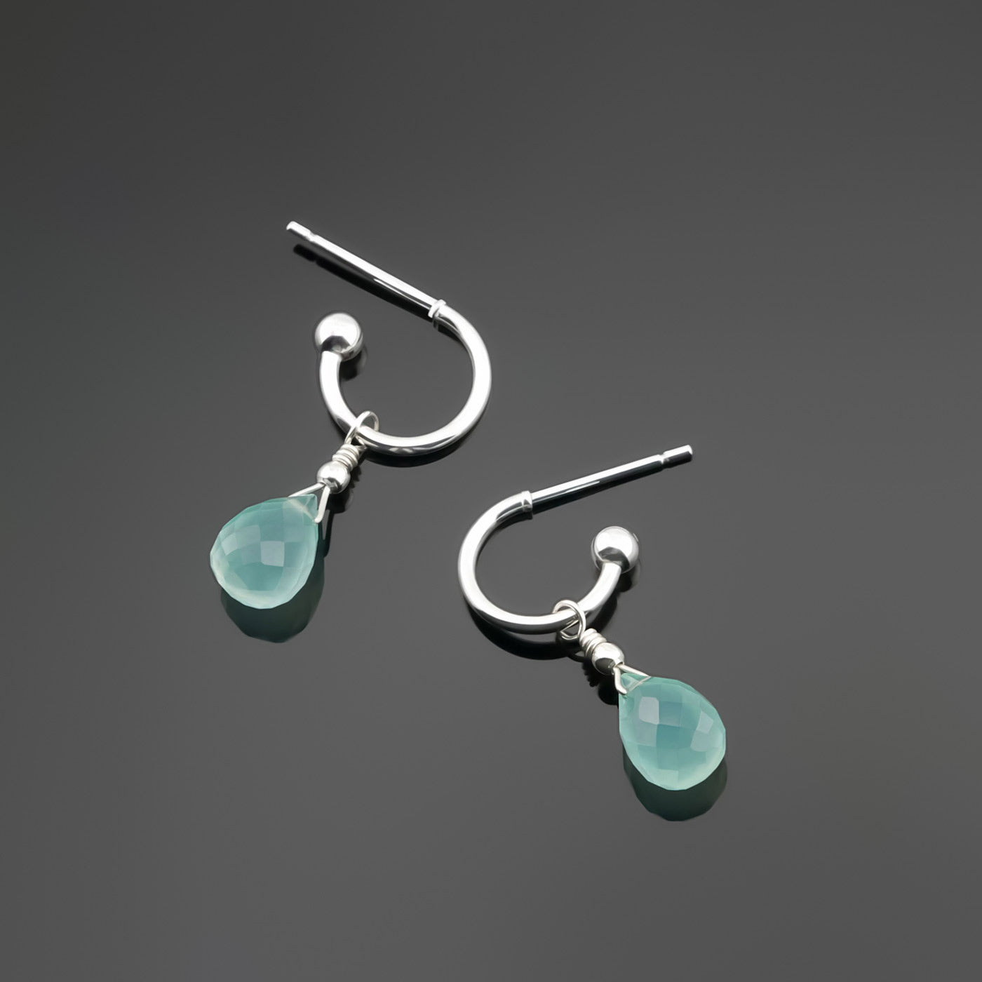 Top view of a pair of mini sterling silver hoop earrings with aqua blue-green teardrop-shaped AAA quality chalcedony gemstones on a dark gray background, top view.