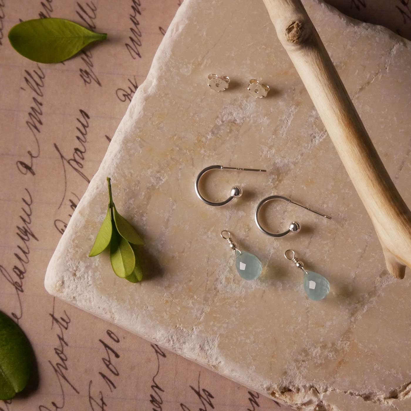 Sterling silver interchangeable mini hoop earrings with blue-green teal AAA quality aqua chalcedony gemstones, on a textured stone surface with leaves and a branch stylized.