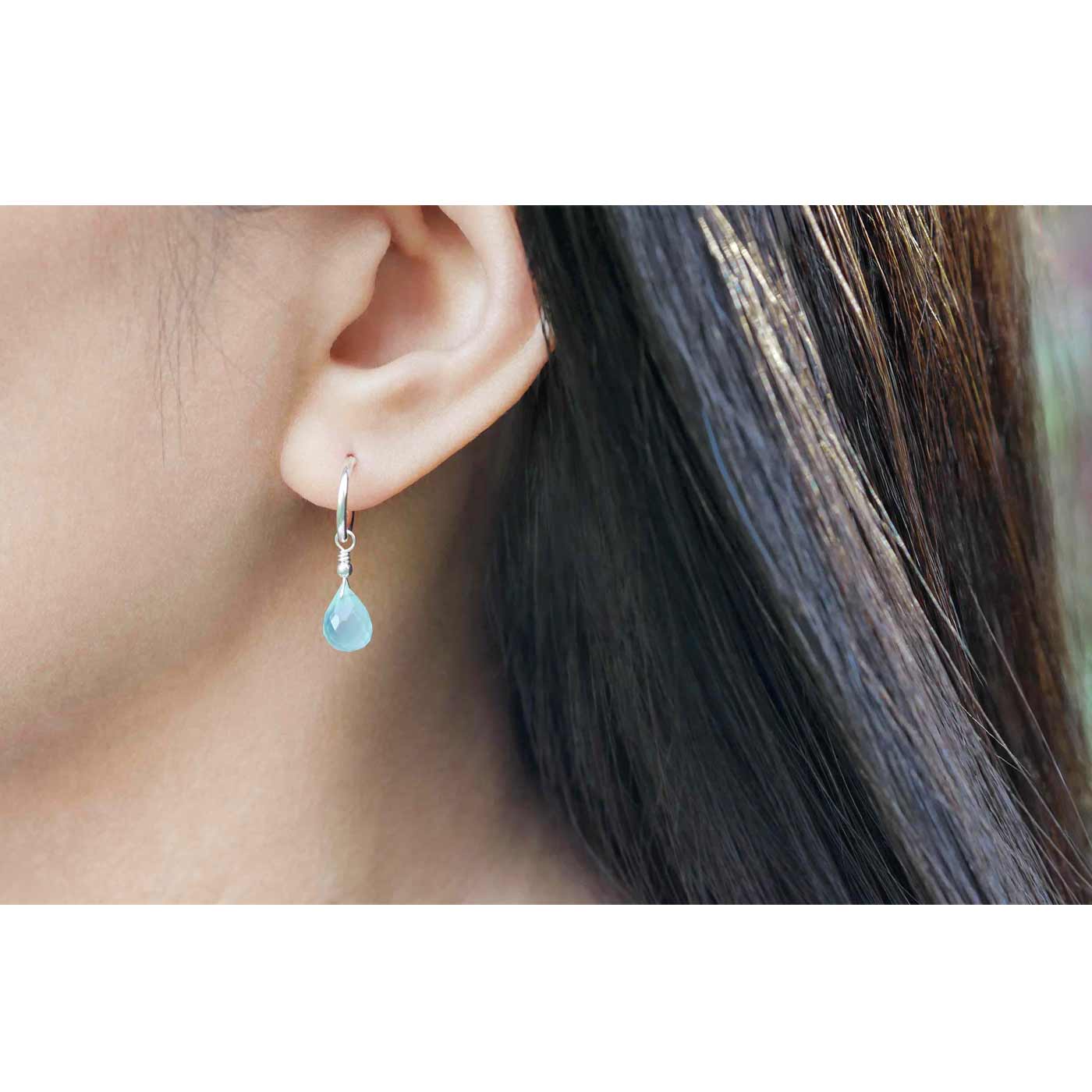 Close-up of a personl's ear wearing  sterling silver interchangeable mini hoop earrings, with blue-green teal AAA quality aqua chalcedony briolette teardrop shaped gemstones with a blurred background.