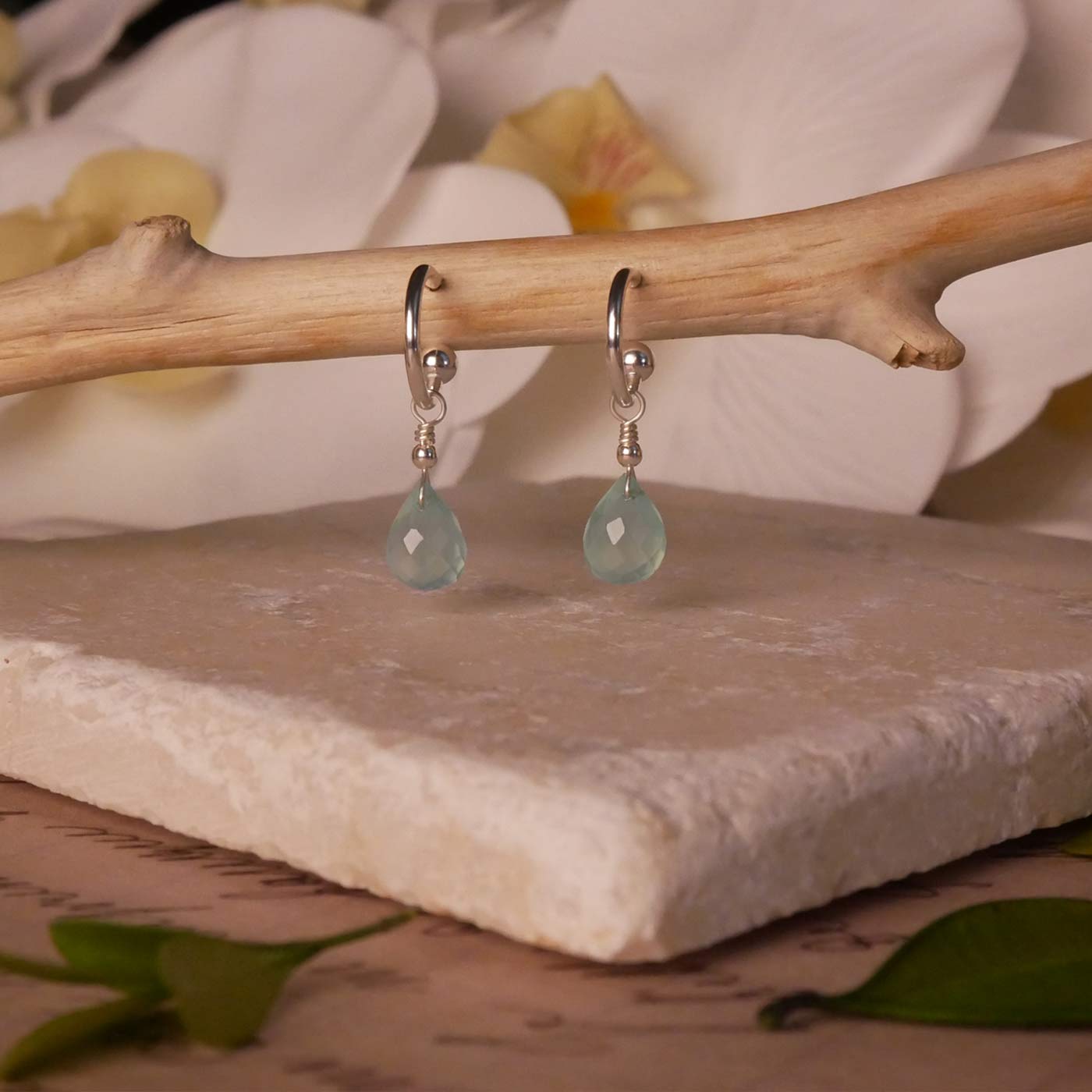 Sterling silver interchangeable mini hoop earrings, with blue-green teal AAA quality aqua chalcedony teardrop briolette shaped gemstones, stylized on a branch, with a few leaves in the foreground, hovering over natural stone, with flowers in the background.