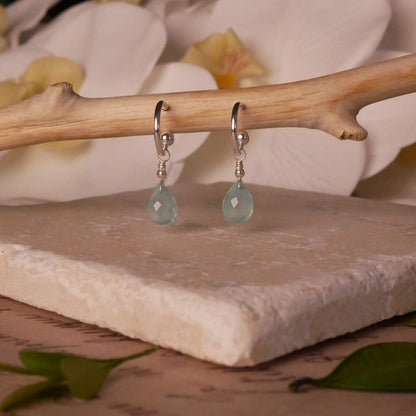 Sterling silver interchangeable mini hoop earrings, with blue-green teal AAA quality aqua chalcedony teardrop briolette shaped gemstones, stylized on a branch, with a few leaves in the foreground, hovering over natural stone, with flowers in the background.