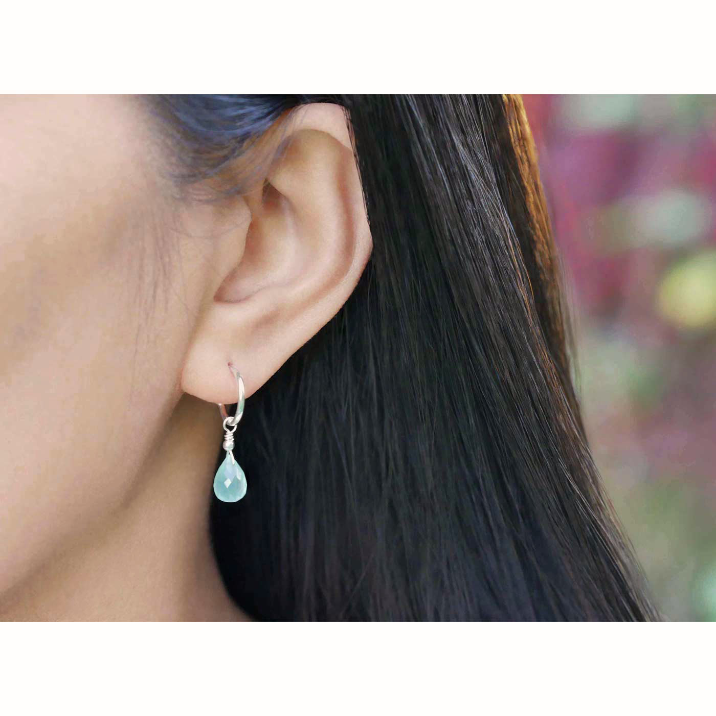 Close-up of a person's ear wearing sterling silver interchangeable mini hoop earrings, with blue-green teal AAA quality aqua chalcedony teardrop shaped briolette gemstones, with a blurred background, the model has black hair.