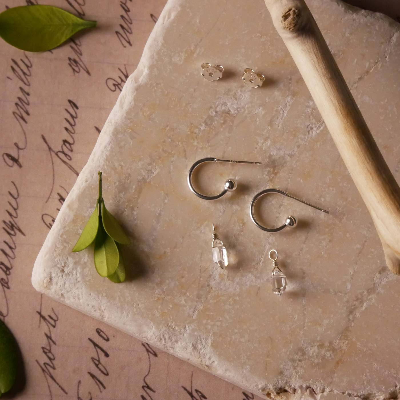 Pair of dainty high quality Herkimer Diamond Quartz drops, with Sterling Silver hoop earrings laying separately to show detachable earrings, laying on a stone surface stylized with green leaves and a tree branch.