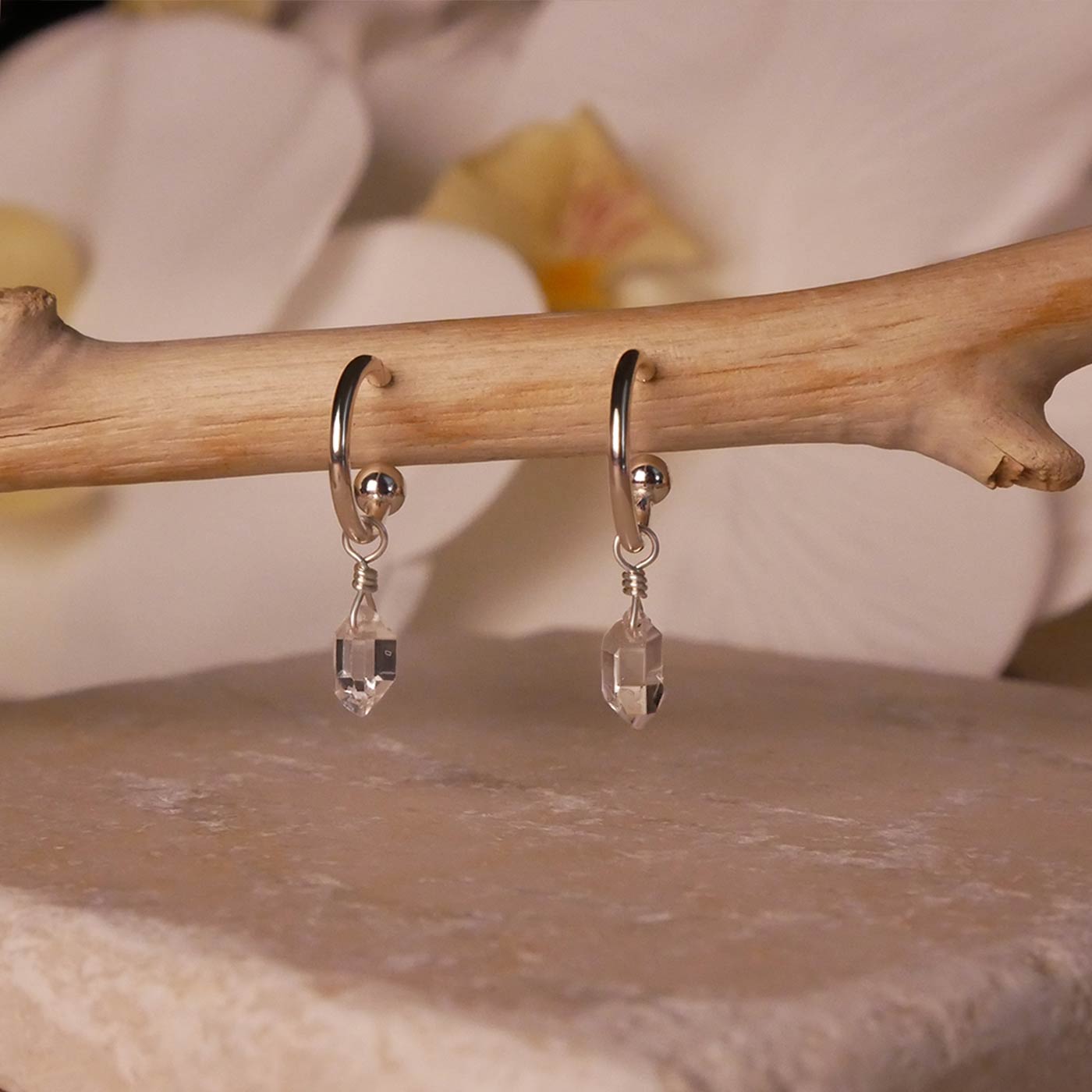 Pair of dainty high quality Herkimer Diamond Quartz drops on Sterling Silver hoop earrings dangling from a tree branch, stylized with a stone surface below, and white flowers blurred in the background.
