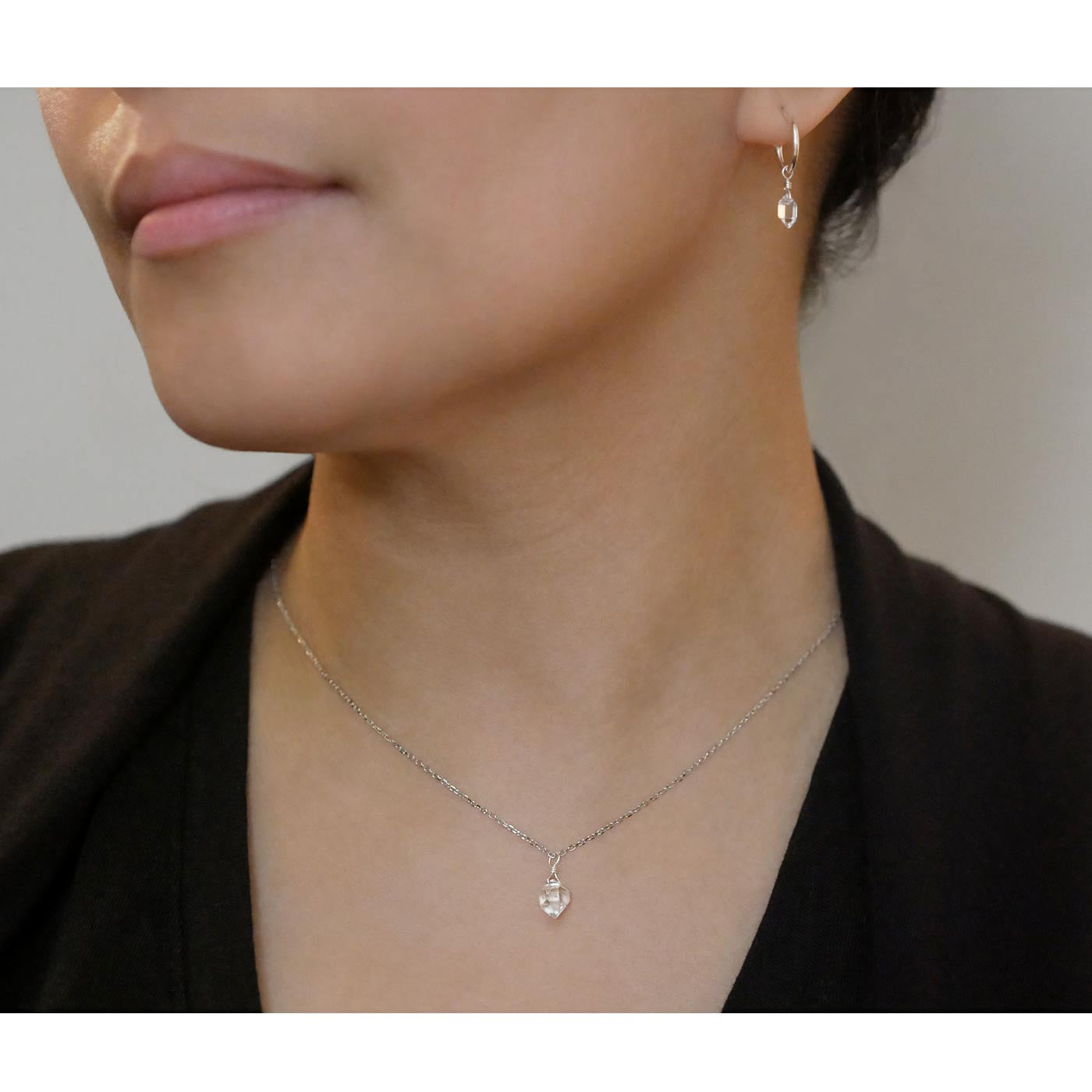Pair of dainty high quality Herkimer Diamond Quartz drops on Sterling Silver hoop earrings and a dainty Herkimer Diamond Quartz drop pendant necklace, worn by a person with their hair up and wearing a black top.