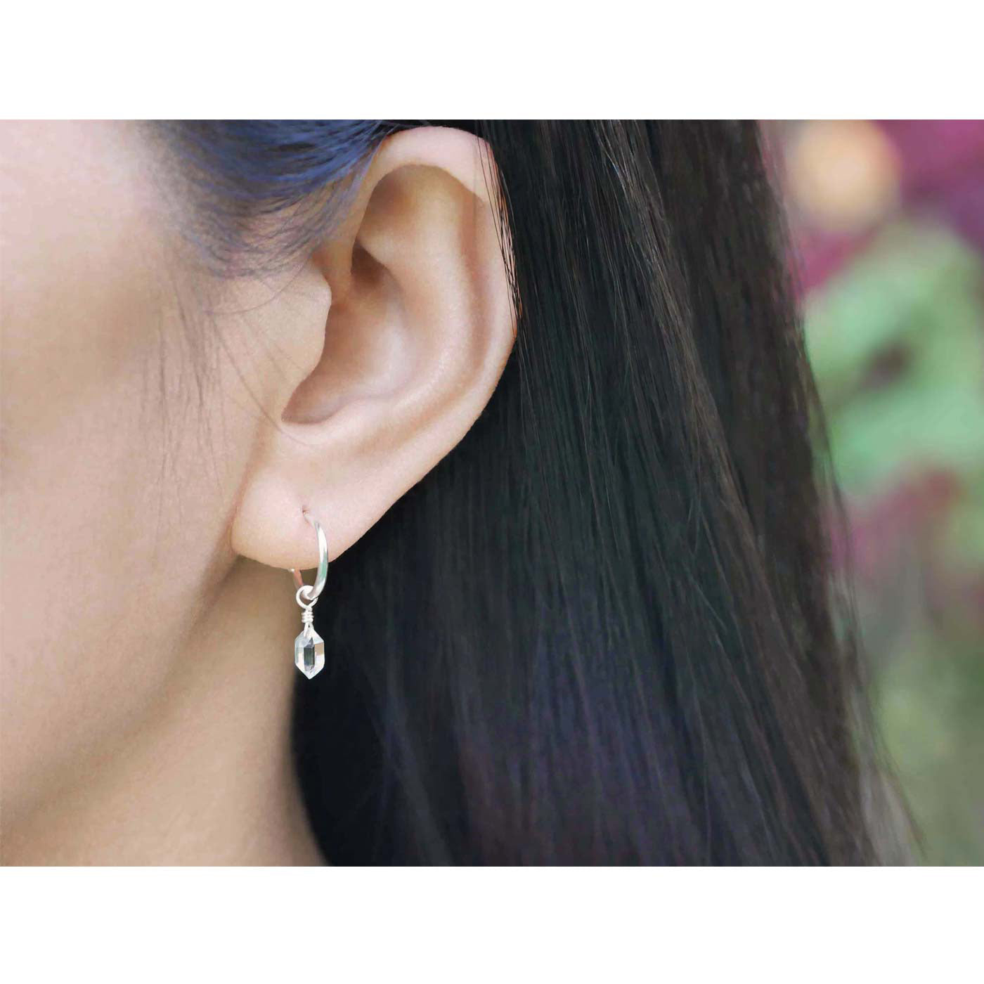 Close up of dainty, high quality Herkimer Diamond Quartz drop on Sterling Silver hoop earring, worn outside by a person with long, dark hair and plants and flowers blurred in the background.