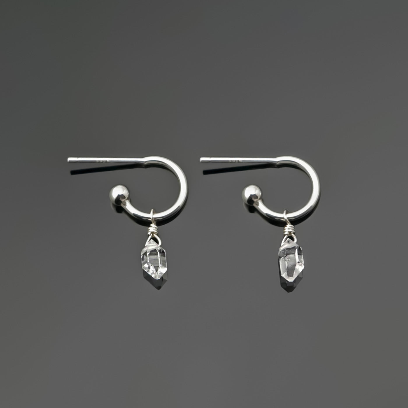 Pair of dainty high quality Herkimer Diamond Quartz drops on Sterling Silver hoop earrings laying on a dark gray background, top view.