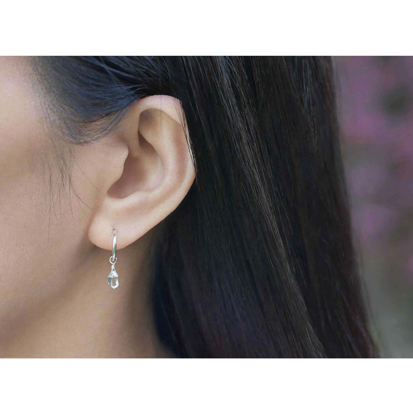 Dainty, high quality Herkimer Diamond Quartz drop on Sterling Silver hoop earring, worn outside by a person with long, dark hair and plants and flowers blurred in the background.