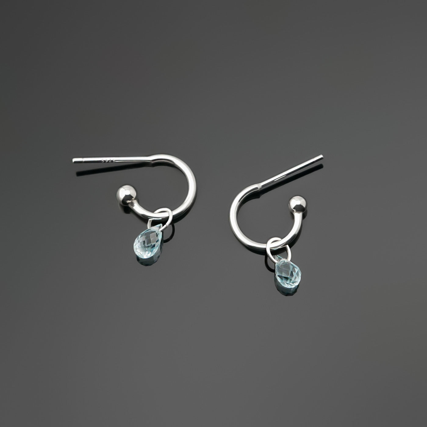 Pair of blue topaz gemstone silver hoop earrings on a dark gray background, top view.