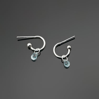 Pair of blue topaz gemstone silver hoop earrings on a dark gray background, top view.