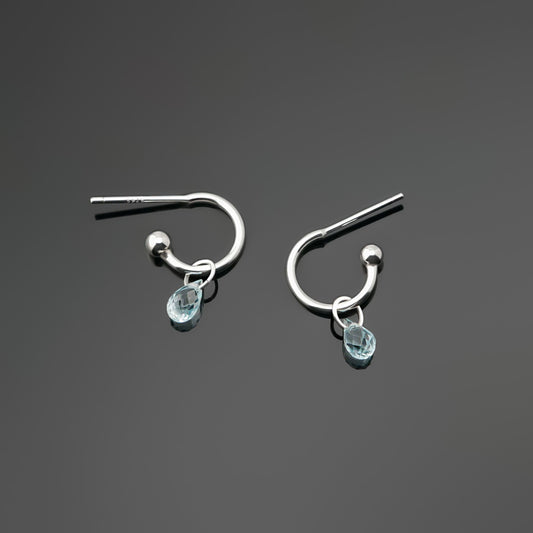Pair of blue topaz gemstone silver hoop earrings on a dark gray background, top view.