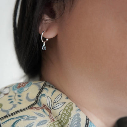Close-up of a blue topaz gemstone silver hoop earring, worn on a model's ear with a blurred background.