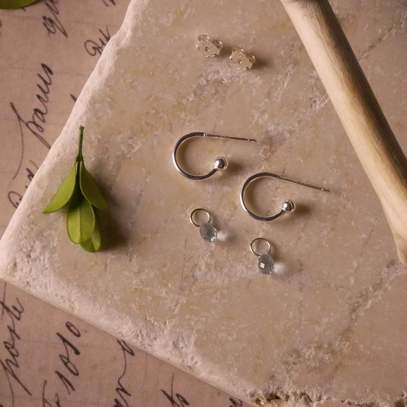 Pair of blue topaz gemstone silver interchangeable hoop earrings, stylized on a textured stone surface with a branch and green leaves. The blue topaz charms are laying separately.