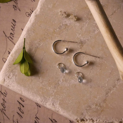 Pair of blue topaz gemstone silver interchangeable hoop earrings, stylized on a textured stone surface with a branch and green leaves. The blue topaz charms are laying separately.