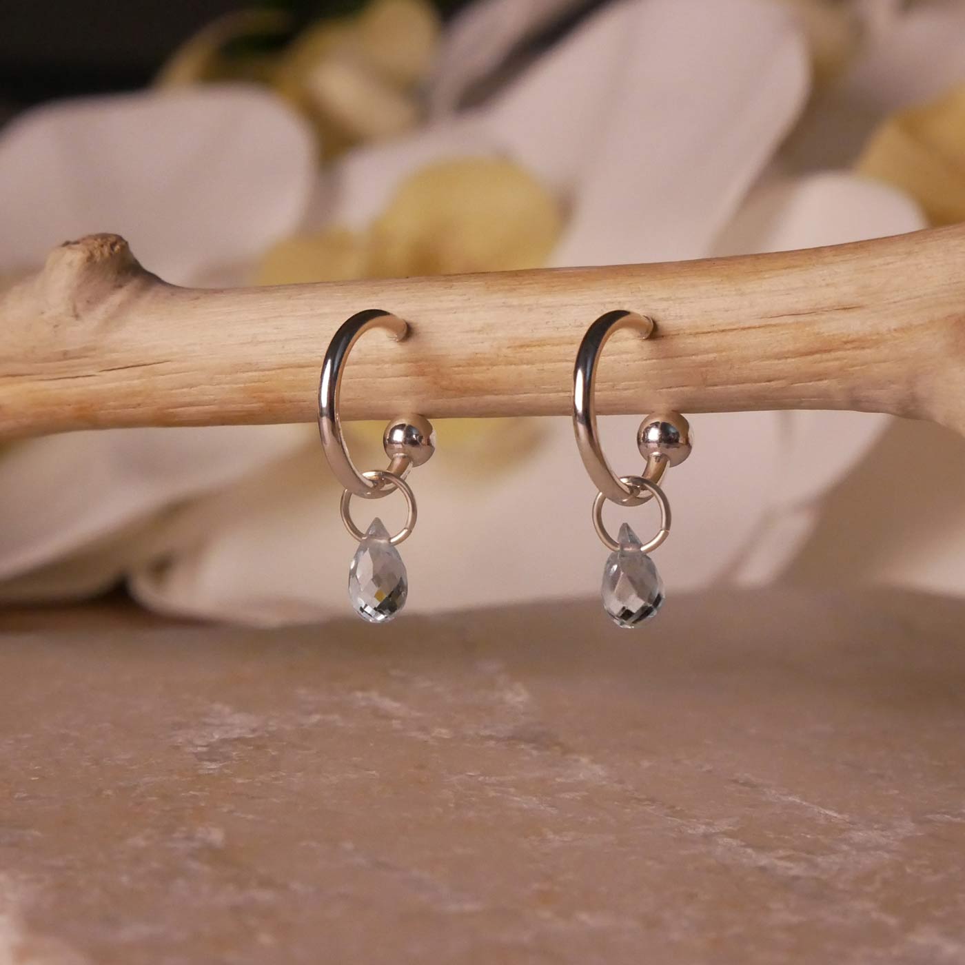 Pair of dainty blue topaz gemstone sterling silver interchangeable hoop earrings, hanging on a wooden branch. The background is stylized with stone underneath and flowers blurred in the background.