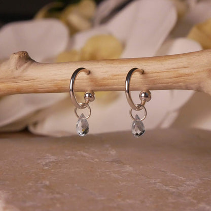 Pair of dainty blue topaz gemstone sterling silver interchangeable hoop earrings, hanging on a wooden branch. The background is stylized with stone underneath and flowers blurred in the background.