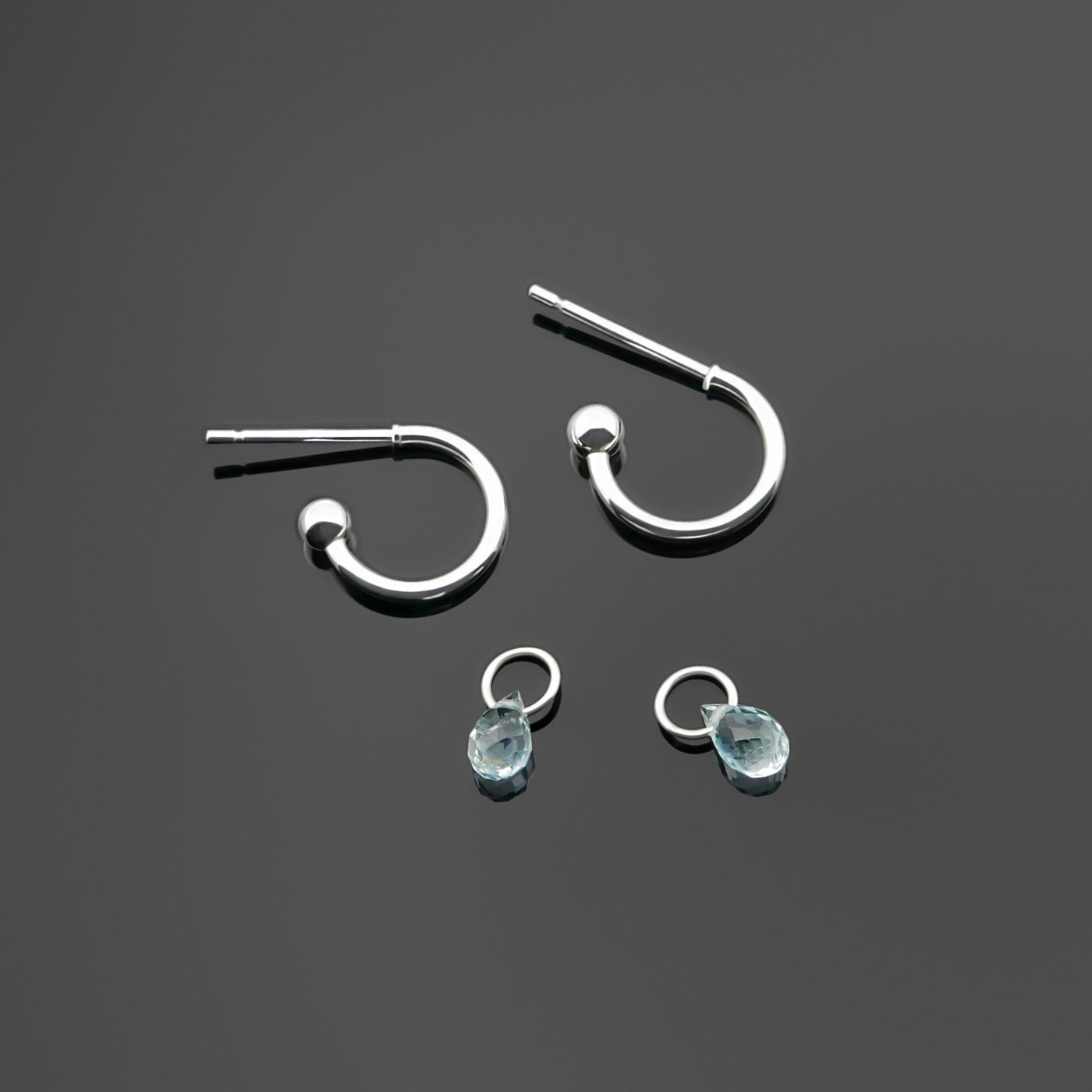 Pair of blue topaz gemstone silver interchangeable hoop earrings, with the blue topaz drop charms laying separately. It is a top view on a plain dark gray background.