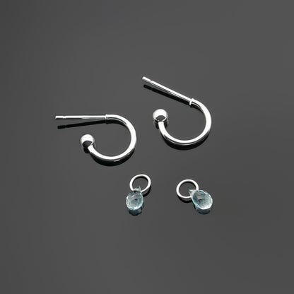 Pair of blue topaz gemstone silver interchangeable hoop earrings, with the blue topaz drop charms laying separately. It is a top view on a plain dark gray background.