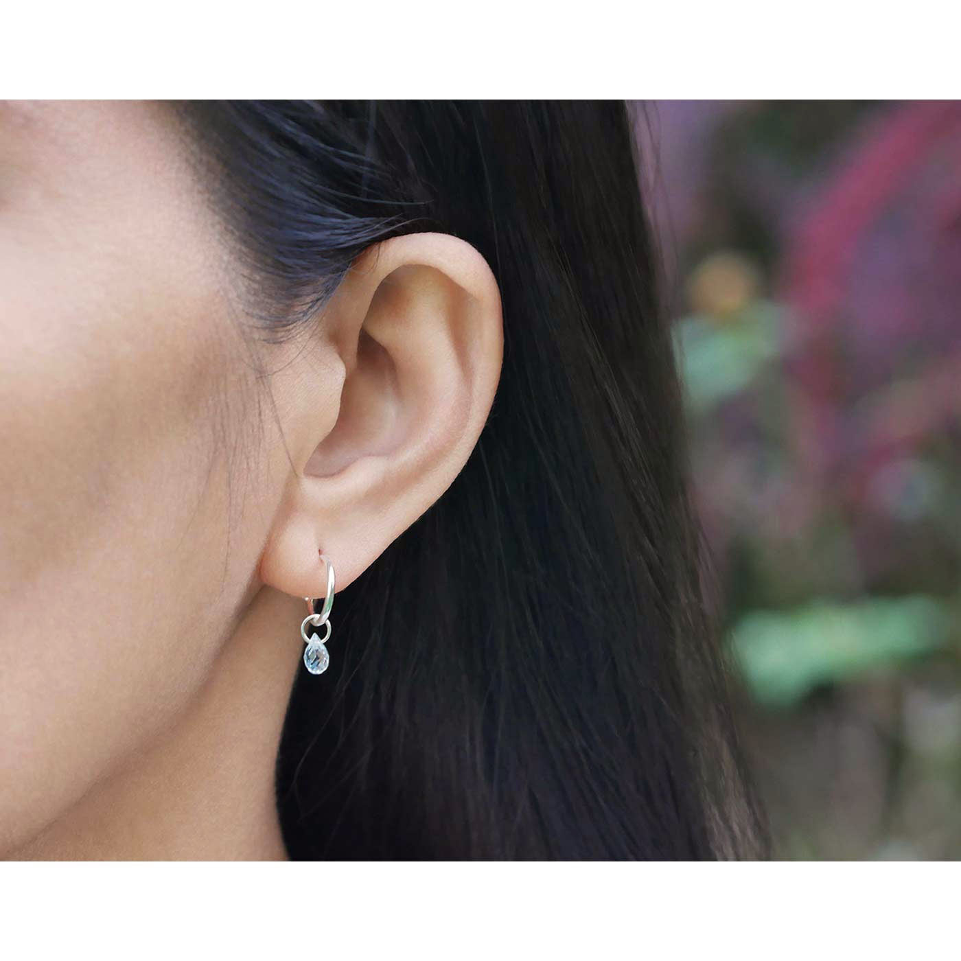 Close-up of a model's ear wearing a blue topaz gemstone sterling silver interchangeable hoop earring. The setting is outside, the background has plants and flowers blurred, the model has black hair.