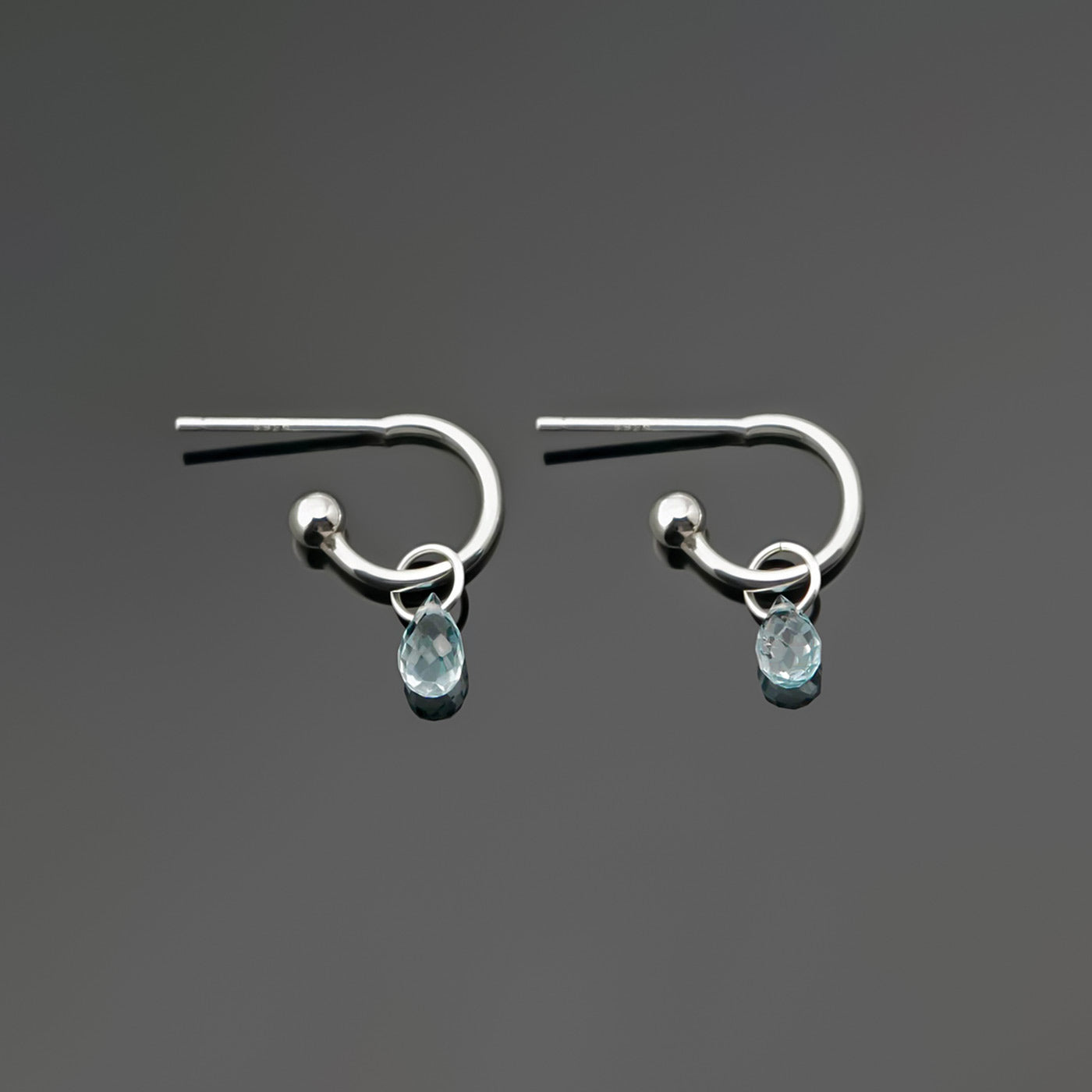 A pair of blue topaz gemstone sterling silver interchangeable hoop earrings, laying flat on a dark gray background, top view.