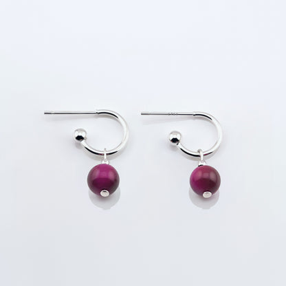 Sterling silver hoop earrings with dark pink tiger's eye round sphere charms laying flat on a plain white background, top view.