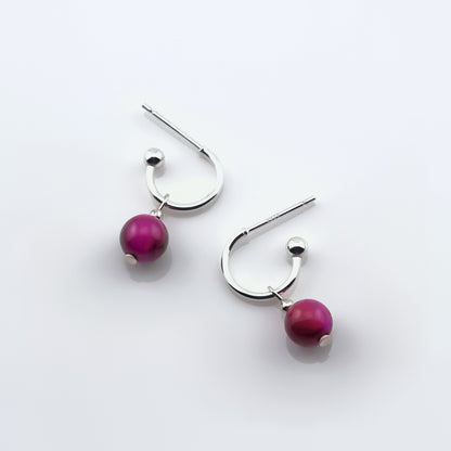 Sterling silver hoop earrings with dark pink tiger's eye round sphere charms laying flat on a plain white background, top view.