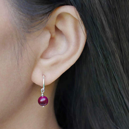 Close-up of a person's ear wearing a sterling silver hoop earring with a dark pink tiger's eye round sphere charm. Person has long black hair.
