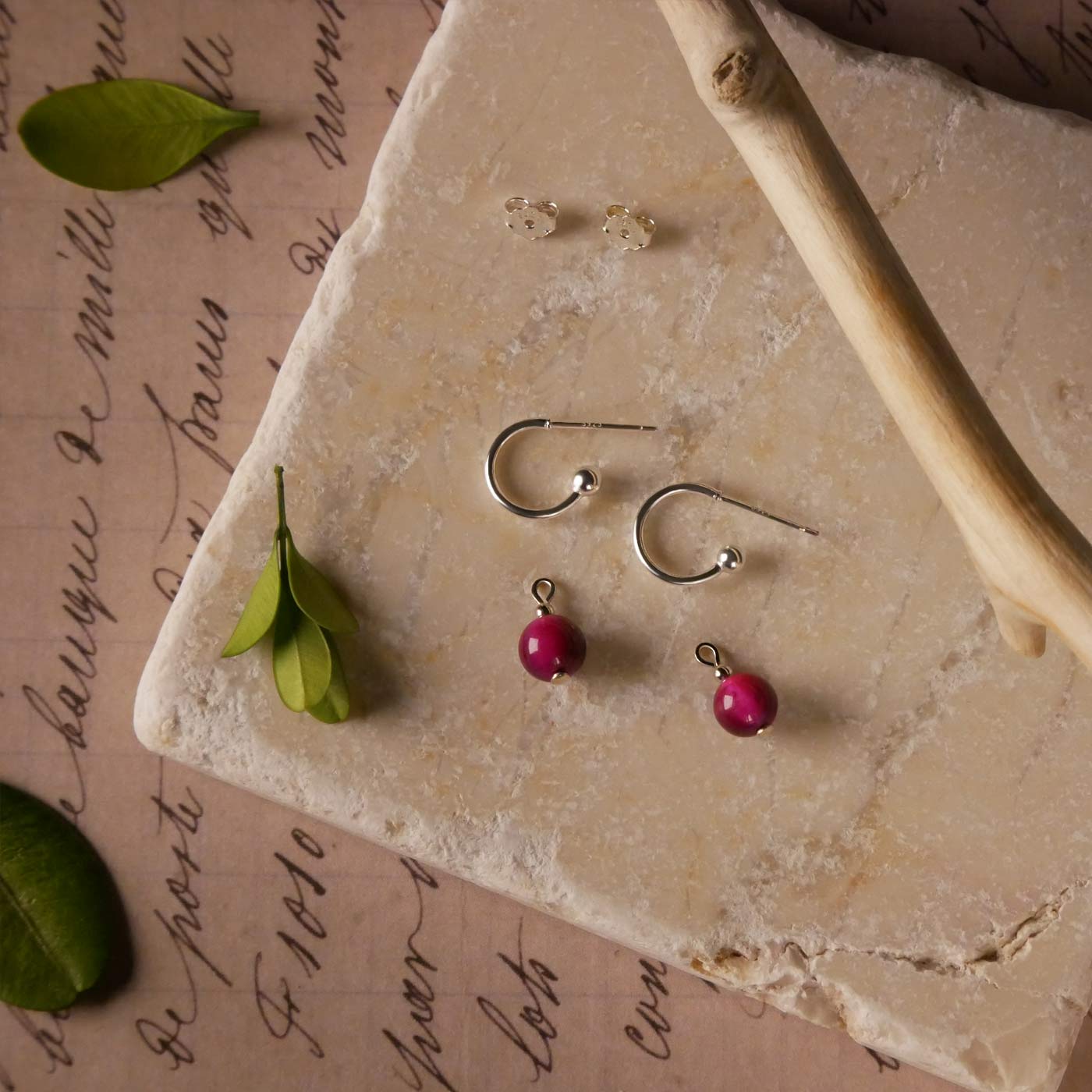 Sterling silver hoop earrings with dark pink tiger's eye round sphere charms laying flat on a stylized background. They are laying on a textured stone surface, green leaves, a tree branch, and vintage paper with  cursive writing underneath.