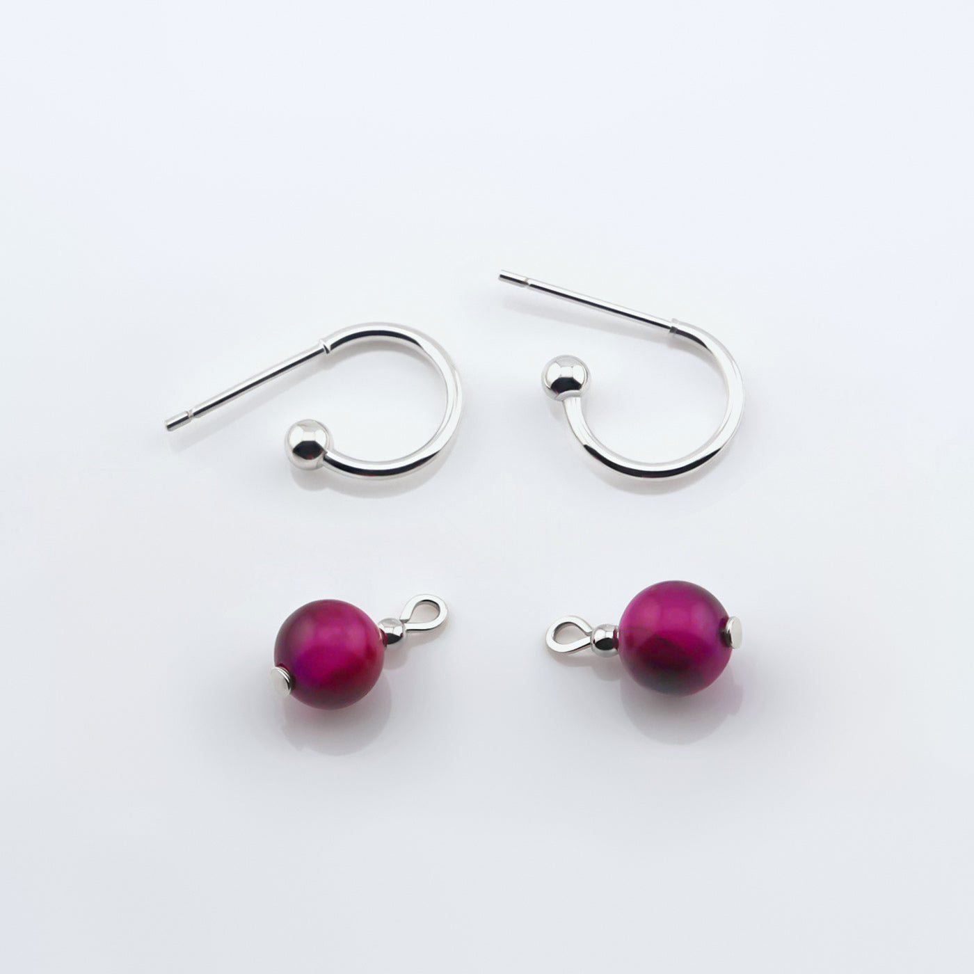 Sterling silver hoop earrings with interchangeable dark pink tiger's eye round sphere charms laying flat on a plain white background, top view.
